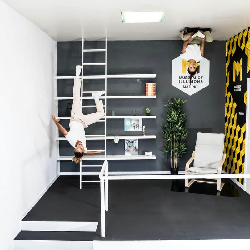 Museum of Illusions Madrid City Card (Madrid) Two people appear upside down in an optical illusion room at the Museum of Illusions Madrid, with furniture and shelves on the walls.