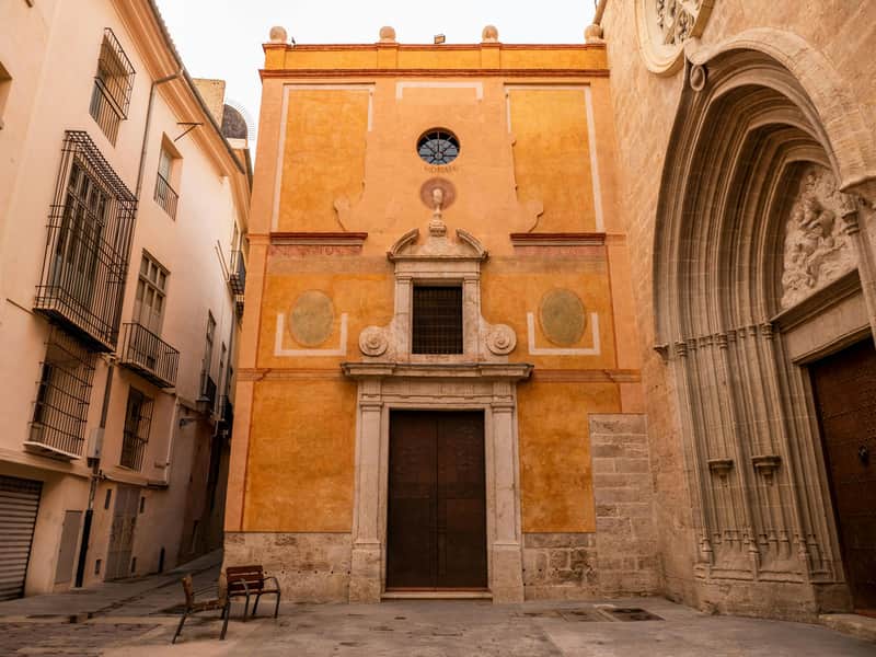 Church of San Nicolás Valencia City Card (València) A narrow courtyard with benches, surrounded by beige and yellow historic buildings with ornate doors and barred windows.