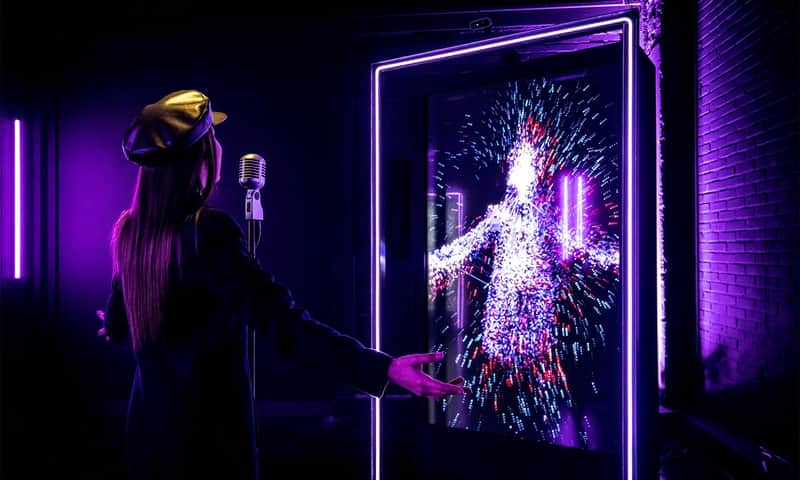 A person in a dark outfit and hat sings into a retro microphone. A vibrant digital display shows a colorful, abstract figure.