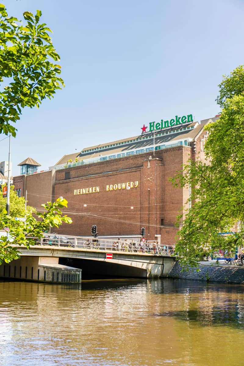 A brick building with "Heineken Brouwerij" sign, next to a bridge over a canal, framed by green trees under a clear blue sky.