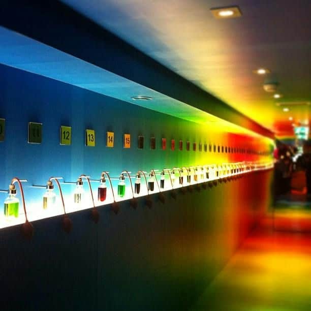 A long hallway with numbered shelves displaying glass containers filled with colored liquids, illuminated by vibrant, colorful lighting.