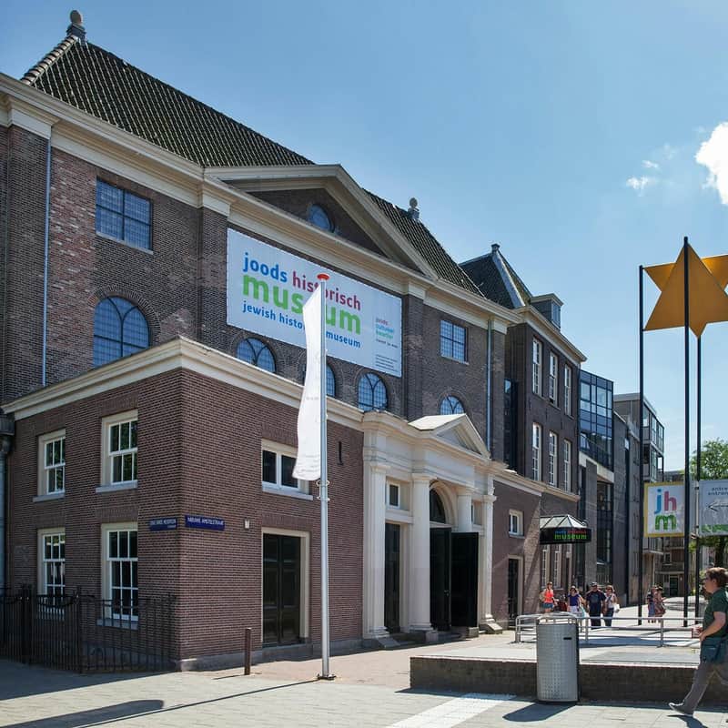 Red-brick building with a sign reading "Jewish Historical Museum" in front, people walking around, and a blue sky above.