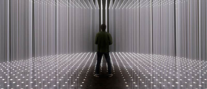 A person stands in a room with vertical light bars on walls and ceiling, and a floor of small, evenly spaced lights.