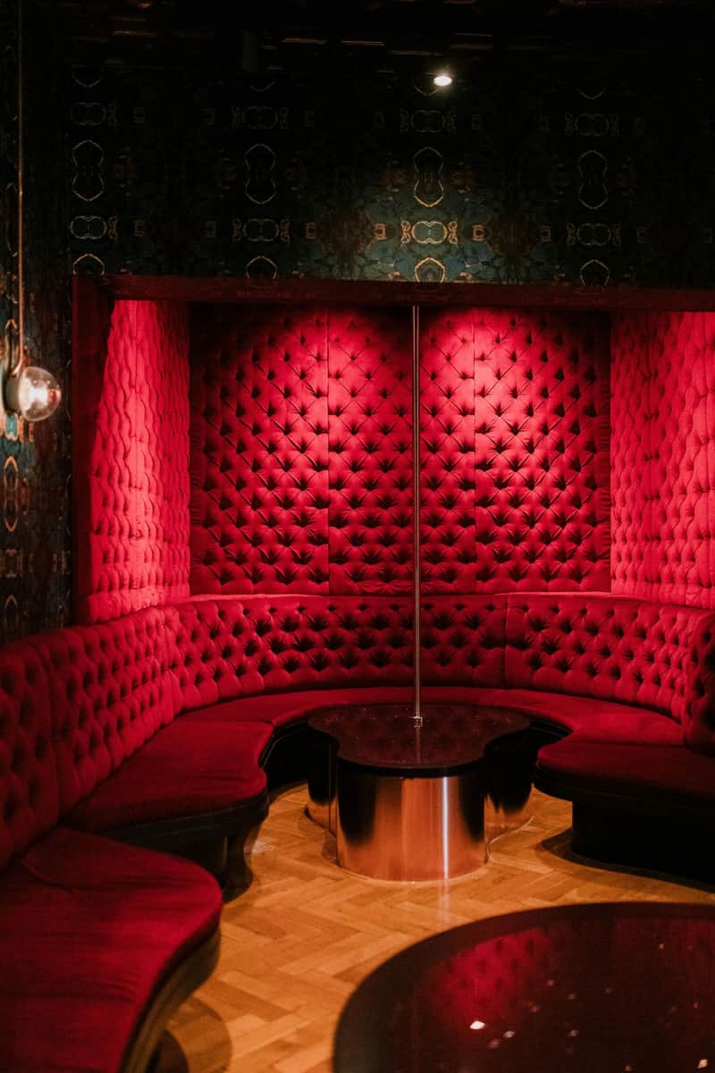 A dimly lit room with red, tufted velvet seating arranged in a semicircle around a pole. There's a round black table in the center.