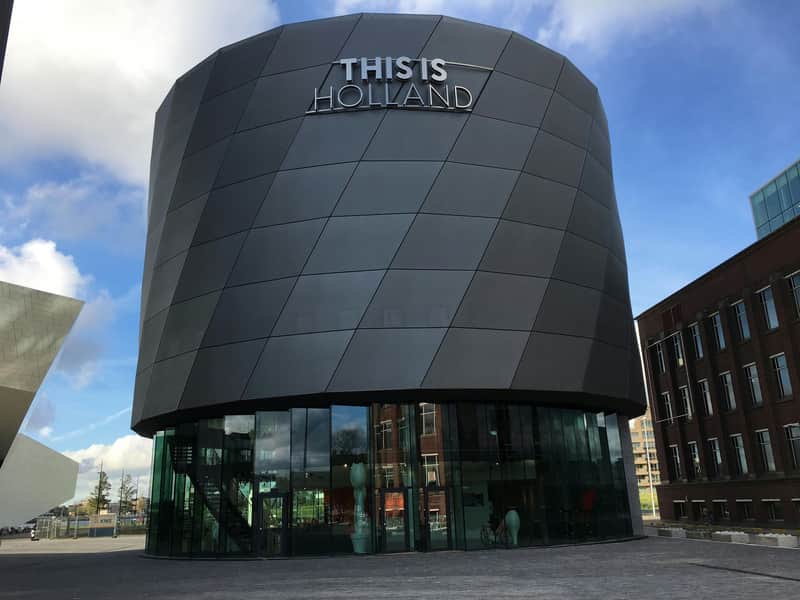 Round, modern building with "THIS IS HOLLAND" sign against a blue sky, with nearby structures and glass entrance at ground level.