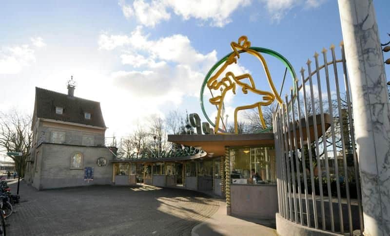 Entrance gate with decorative sculpture and a ticket booth to the right. A brick building and bicycles are visible to the left.