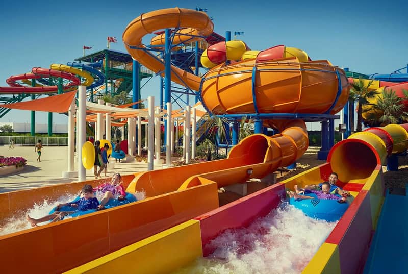 People splashing down colorful water slides on inflatable tubes at an outdoor water park under a clear blue sky.