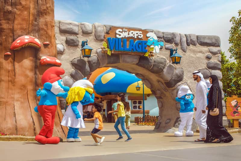 People interact with Smurf characters near the entrance of "The Smurfs Village" theme park, adorned with colorful mushroom houses.