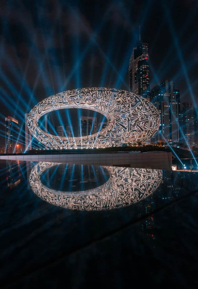 Museum of the Future City Card (Dubai) A futuristic oval-shaped building with glowing script patterns, set against a cityscape with skyscrapers and illuminated by blue beams.