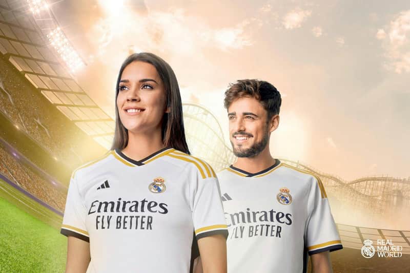 A woman and a man smiling, wearing Real Madrid jerseys, in a brightly lit stadium with a sunset sky. Real Madrid World logo in the corner.