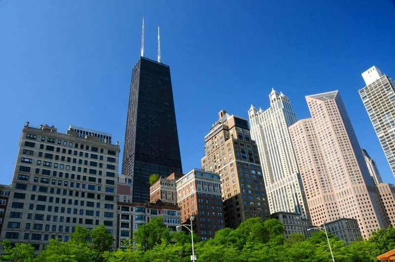 A skyline with various high-rise buildings against a clear blue sky, with trees in the foreground.