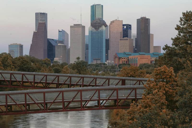 City skyline with tall buildings and a mix of architectural designs. Trees and two metal bridges over a river fill the foreground.
