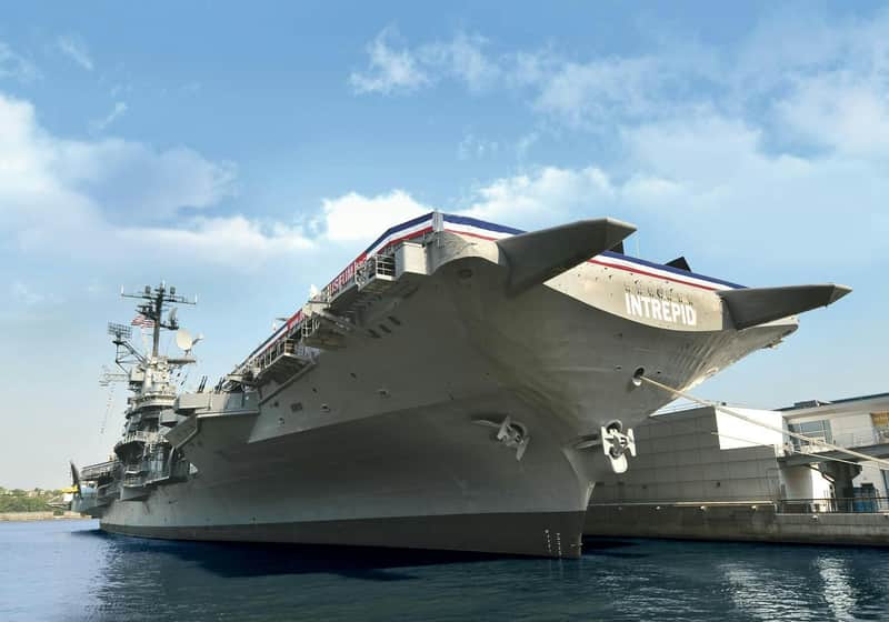 Intrepid Museum City Card (New York) Large aircraft carrier Intrepid docked at a pier, viewed from the bow, under a partly cloudy sky.