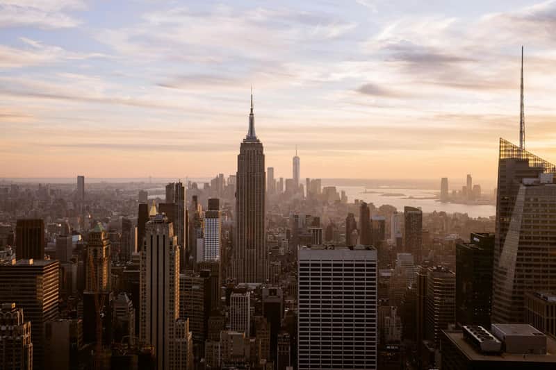 Top of the Rock City Cards & Passes (New York) View of a city skyline at sunset, dominated by tall skyscrapers, with a river and distant buildings in the background.