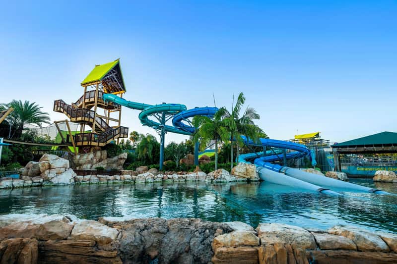 Aquatica Orlando Bykort (Orlando) Et tropisk vandland med en stor, snoet blå vandrutsjebane, palmer, klipper og en pool under en klar blå himmel.