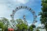 A large Ferris wheel with red gondolas, surrounded by trees, under a partly cloudy blue sky.