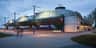 A modern building with large windows and industrial design features, under an evening sky with streetlights and silhouettes of people.