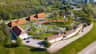Aerial view of a park with pathways, playgrounds, brown-roofed buildings, parking lot, and surrounding greenery on a sunny day.