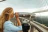 A person with long hair looks through a coin-operated binocular viewer on an outdoor observation deck.