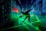 A person in a dark room navigates through green laser beams, with digital graphics and neon lights on the walls.