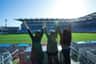 Three individuals with raised arms stand facing a large, empty stadium under bright sunlight.