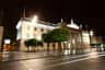 Illuminated neoclassical building with columns and statues at night, surrounded by trees and a wet pavement reflecting lights.