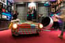 Museum exhibit with a colorful car, posters on the walls, and visitors interacting with displays and taking photos.