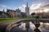 A historic stone cathedral with a tall spire beside a reflective fountain, surrounded by green lawns and a few people under a blue sky.