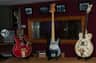 Three guitars on stands in a music studio, along with a reel-to-reel tape recorder. Framed photos hang on a burgundy wall.