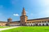 Historic brick castle with a tall central tower, arched windows, and a green lawn, against a clear blue sky with a few clouds.