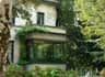 A house with large glass windows, covered in climbing plants and surrounded by lush greenery and trees.