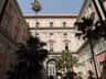 Sunlit courtyard with pink and cream buildings, arched windows, and tall palm trees reaching towards a clear blue sky.