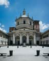 A historic building with a dome, arches, and a statue in the front courtyard, surrounded by columns and a clear blue sky.