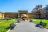 Tourists relax on benches in a courtyard with a large bronze sculpture and historic building under a clear blue sky.