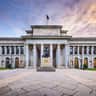 Grand neoclassical building with a statue in front, columns, sculptures, a relief, and a flag atop, under a colorful sky.