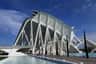 A futuristic white building with structural arches, surrounded by water and a few tall, narrow trees against a blue sky.