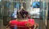 A royal crown with jewels displayed on a red velvet cushion inside a glass case, surrounded by reflective surfaces and lights.