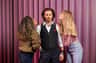 Three people pose in front of a pink curtain; a man in a vest stands between two women, one on each side, both leaning in toward him.