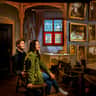 A couple seated on a wooden bench in a room decorated with ornate paintings, a window, and wooden furniture.