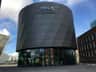 Round, modern building with "THIS IS HOLLAND" sign against a blue sky, with nearby structures and glass entrance at ground level.