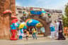 People interact with Smurf characters near the entrance of "The Smurfs Village" theme park, adorned with colorful mushroom houses.