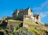 Historic stone castle on a rocky hilltop under a clear blue sky, featuring multiple connected buildings and sloped grassy areas.