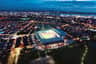 Vista aèria d'un estadi ben il·luminat enmig de les llums de la ciutat al capvespre, amb zones residencials i urbanes al voltant.