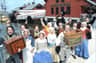 A group in historical attire cheers, holding boxes in front of the Boston Tea Party Ships & Museum by the water.
