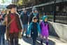 A person in historical attire leads a group of people, including children in colorful winter coats, walking on a sidewalk.