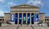 Large, classical-style building with columns and banners promoting "Máximo The Titanosaur" exhibit. People sit on the steps.