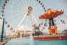 A Ferris wheel and a carousel with swings located near a pool with people around, all under a clear sky at Navy Pier.