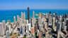 Aerial view of a city with numerous tall buildings near a large body of water under a clear blue sky.