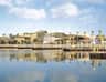 A waterside view of a modern building with palm trees, docks, and calm water reflecting the scene under a partly cloudy sky.