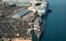 Aerial view of a cruise ship and a battleship docked at a port with adjacent parking lots and terminal buildings.